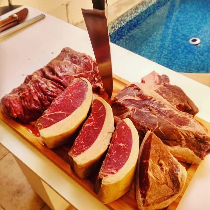 Tábua com cortes de carnes cruas variados e faca, à beira da piscina