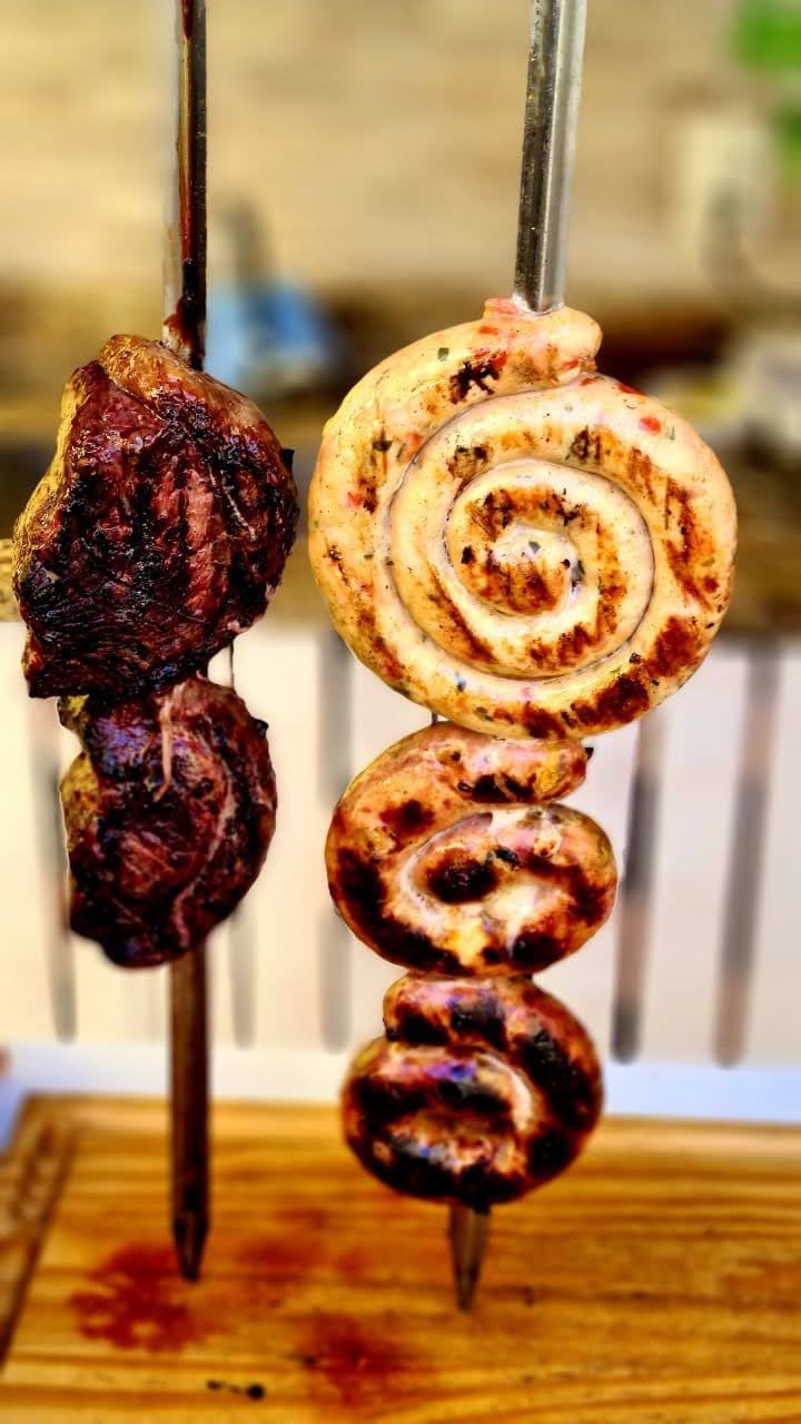 Espeto de carne assada ao lado de linguiça recheada enrolada no espeto